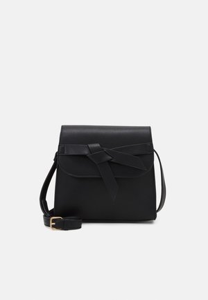 Sort crossbody taske med klap og dekorativt knyttet remdetalje, justerbar skulderrem og guldfarvet spænde på hvid baggrund.