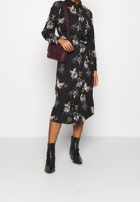 Abito midi nero floreale con maniche lunghe, colletto e cintura da annodare. Abbinato a una borsa a tracolla bordeaux e stivaletti neri.