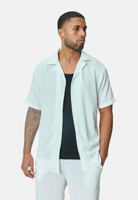 Chemise blanche à manches courtes avec un tissu texturé, portée sur un débardeur noir. Présente un col classique et une coupe ample.