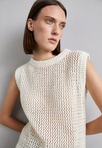 Maglione senza maniche in crema con una lavorazione a maglia morbida e aperta, dotato di un collo a coste e di armhole leggermente allungate.