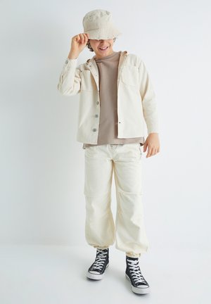 Chapeau seau beige, veste beige avec deux poches, haut marron et pantalon beige clair. Baskets montantes noires avec semelles blanches.