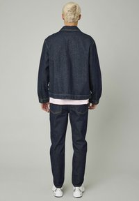 Veste en denim bleu foncé et jeans avec une coupe décontractée. Elle a un col, des poignets boutonnés et des surpiqûres plus claires. Portée avec une chemise rose en dessous.