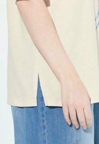 Brazo y mano derechos de una persona que lleva una camisa beige con una abertura lateral y jeans de mezclilla azules.