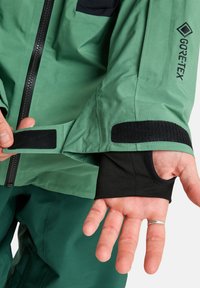 Hand die de manchette van een groene Gore-Tex jas met zwarte klittenbandafsluiting aanpast, draagt een zwarte binnenmouw met duimopening en een zilveren ring om de vinger.