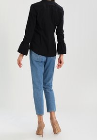 Blouse noire à manches évasées, jean denim bleu clair. Mannequin portant des talons nus, de dos, mettant en valeur les détails de la tenue.