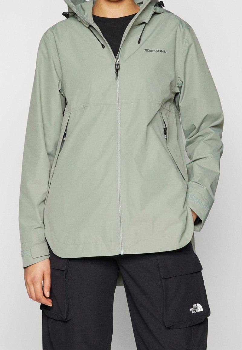 Giacca impermeabile verde chiaro con zip frontale, cappuccio regolabile e tasche laterali. Presenta un logo distintivo sul petto e sui polsini.