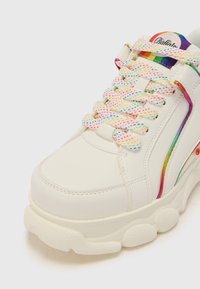 Zapatilla blanca con una suela gruesa y texturizada, que presenta cordones moteados de varios colores y un acento de arcoíris a lo largo del costado.