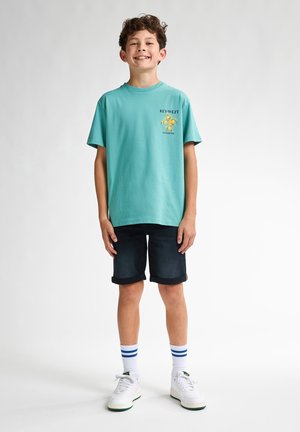 T-shirt en coton turquoise avec un motif de crabe et du texte, associé à un short en jean foncé, des baskets blanches et des chaussettes rayées.