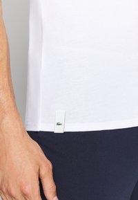 T-shirt in cotone bianco con un piccolo etichetta verde Lacoste sul margine sinistro, abbinata a pantaloni blu navy. Inquadratura ravvicinata della mano e del tessuto.
