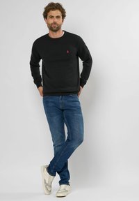Schwarzer Pullover mit gerippten Bündchen und Saum, featuring ein kleines rotes Logo auf der Brust. Getragen mit blauen Jeans und weißen Turnschuhen.