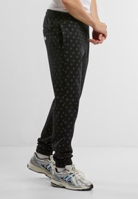 Sorte joggebukser med et subtilt logomønster, elastisk livlinje og ribbestrikkede mansjetter; kombinert med metalliske joggesko med blå detaljer.