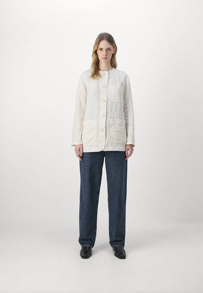 Veste en lin crème à col rond, avec des boutons sur le devant et deux grandes poches avant ; associée à un pantalon en denim noir ample.