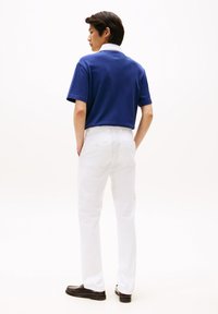 Μπλε ναυτικό πόλο με λευκό γιακά, σετ με λευκό παντελόνι και σκούρα μοκασίνια, φωτογραφημένο από πίσω πάνω σε απλό φόντο.