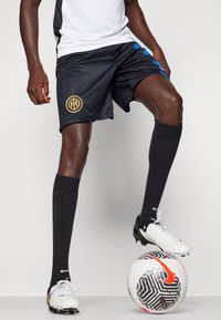 Shorts de sport noirs avec des accents bleus et un logo doré, associés à des chaussettes noires jusqu'aux genoux et des chaussures de football blanches, sur un ballon à motifs.