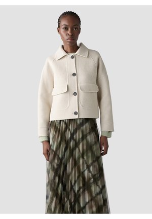 Frau trägt eine cremefarbene Wolljacke mit Knöpfen und Klappentaschen sowie einen grünen, gemusterten, plissierten Rock und steht vor einem einfarbigen Hintergrund.