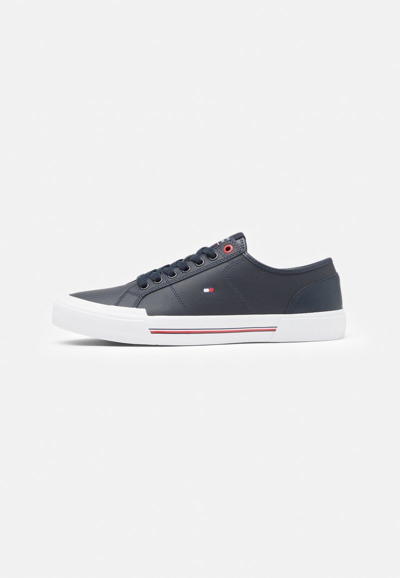 Baskets basses noires avec semelle blanche, lacets noirs, petit logo drapeau sur le côté et détail de rayures rouges et blanches sur la semelle.