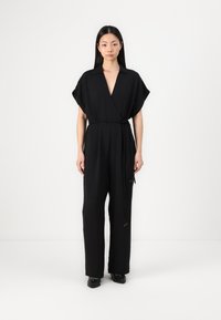 Svart jumpsuit med v-hals og korte ermer, innsnurt i livet med et belte; har teksturert stoff og vide bukser.