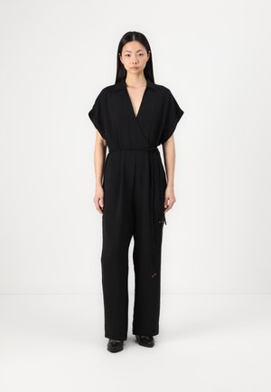 Black jumpsuit with a v-neck and short sleeves, cinched at the waist with a tie; features textured fabric and loose-fitting trousers.