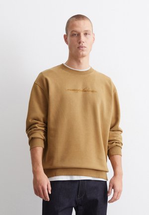Bruine sweatshirt met een ribbelzijde rond de hals en mouwen, voorzien van geborduurde tekst over de borst. Gladde textuur, ontspannen pasvorm, gelaagd over een wit T-shirt.