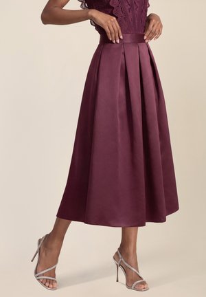 Frau in einem plissierten, hoch taillierten, dunkelvioletten Midi-Rock mit Spitzentop, die silberne Riemchensandalen mit hohen Absätzen trägt, steht vor einem einfarbigen Hintergrund.