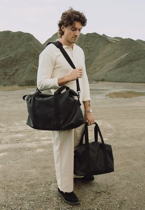 Ung mann i hvit antrekk står ute og holder to svarte duffelbagger foran en bakgrunn av store jordhauger.