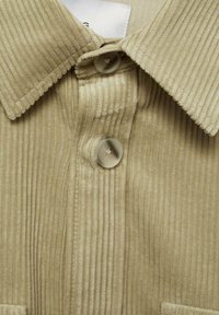 Lichtbeige corduroy shirt met een gestructureerd, ribbelpatroon. Heeft een puntkraag en twee knopen op de voorkant van de sluiting.
