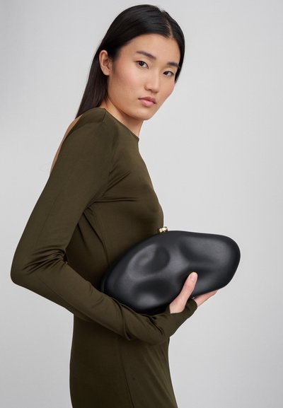 Mustakasi, orgaanisessa, veistoksellisessa muodossa, matta pinta ja kultainen lukko, pidetty oliivinvihreää kangasta vasten.