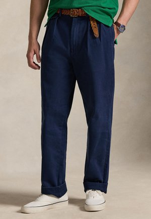 Bărbat purtând pantaloni bleumarin cu tivuri întoarse, adidași albi, o cămașă verde, o curea maro împletită și un ceas de mână.