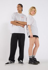 Bílé oversize trička s červeným logem, kombinované s černými kalhotami a botami. Jeden model má na sobě černé kraťasy a bojové boty, druhý má na sobě tenisky.