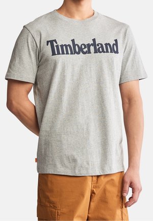Mand iført en lysegrå Timberland t-shirt med korte ærmer og brune cargoshorts, stående foran en ensfarvet baggrund.