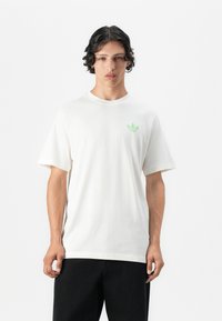 White cotton t-shirt with short sleeves and a crew neck, featuring a small green leaf logo on the left chest. Dark pants are worn.