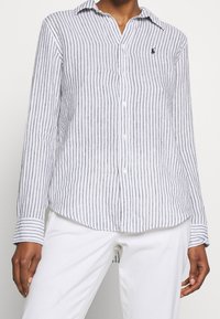 Person klädd i en vit långärmad skjorta med svarta vertikala ränder och vita byxor, stående mot en enkel bakgrund.