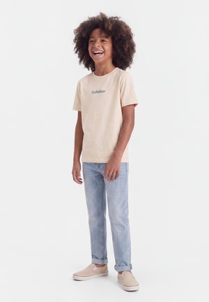Kind mit lockigem Haar, das ein beiges Quiksilver-T-Shirt, hellblaue Jeans und beige Slip-On-Schuhe trägt, lächelnd und vor einem weißen Hintergrund steht.