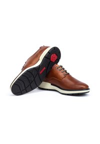 Zapatos de cuero marrón con textura perforada, entresuela blanca y suela de goma negra. Presentan acentos rojos redondos en la suela.