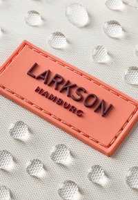 Textúrovaná svetlosivá tkanina s vystupujúcimi kvapkami; koralová etiketa s vyrazeným "LARKSON HAMBURG" v tmavofialovej.