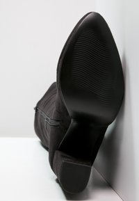 Černé semišové kotníkové boty s texturovanou černou gumovou podrážkou. Detail na bočním zipu a tvarovaný podpatek, který zdůrazňuje hladký povrch a čisté linie.