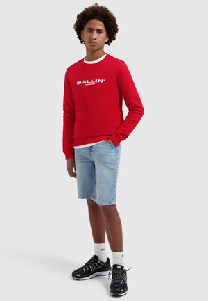 Tiener met krullend haar die een rode "BALLIN" trui draagt, lichte denimshorts, witte Nike sokken en zwarte Nike sneakers, staand met één hand in de zak.