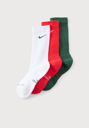 Trois chaussettes côtelées à tige mi-haute, blanches, rouges et vert foncé, chacune ornée d’un petit logo Nike Swoosh, disposées sur un fond clair.