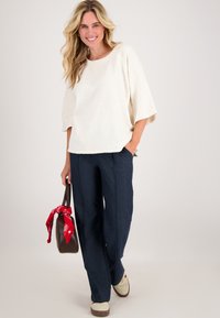 Crème top met korte mouwen en een relaxed fit; gecombineerd met donkerblauwe wijde broek. Houdt een bruine tas vast met een rode bandana om het handvat geknoopt.