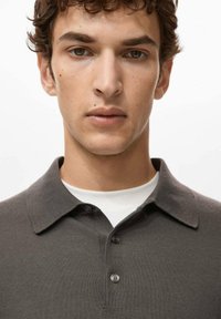 Szara koszulka polo o prążkowanej strukturze, z krótkimi rękawami i zapinanym na guziki karczkiem, noszona na białej koszulce z okrągłym dekoltem.