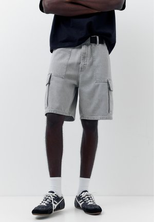 Homme portant un short cargo gris clair, une ceinture noire, des baskets noires avec des lacets blancs, des chaussettes blanches et une chemise foncée, les bras croisés.