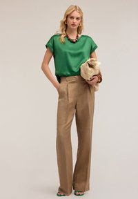 Grön satin topp, beige hög midja byxor med frontveck, håller en beige väska, gröna sandaler med remmar, och ett brunt pärlhalsband.