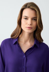 Chemise boutonnée violette avec col, en tissu lisse, offrant une coupe décontractée et un léger éclat, sans motifs ni accents visibles.