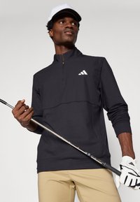 Čierny pulóver s krátkym zipsom a textúrovaným dizajnom, bielym logom na hrudi a rebrovanými manžetami; nosený s béžovými nohavicami a držaním golfovej palice.