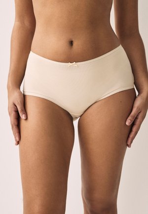 Naine kannab beeži värvi kõrge vöökohaga õmblusteta aluspesu väikese paelaga detailiga, seisab käed lõdvestunult külgedel.