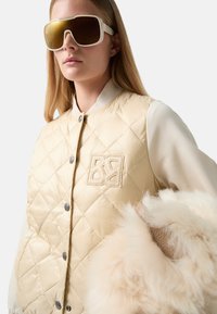 Beżowa pikowana kamizelka z logo, szare guziki i kremowy rękaw. Duże okulary słoneczne filtrujące światło oraz puszysty biały detal akcentowy.