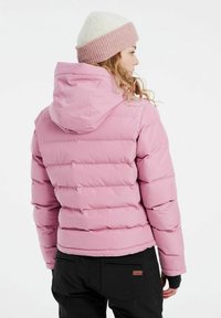 Rosa Pufferjacke mit Kapuze, gestepptem Design und gerippten Bündchen. Getragen mit einer creme-rosa gestrickten Mütze, ergänzt durch schwarze Hosen.