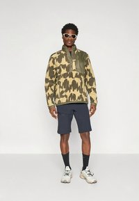 Žalios ir smėlio spalvos kamufliažinis megztinis su užtrauktuku ant apykaklės ir priekinė kišene, derinamas su tamsiai mėlynais šortais ir lengvais sportiniais bateliais.