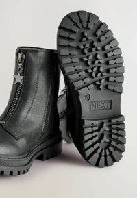 Bottines en cuir noir avec semelles épaisses, fermeture éclair sur le côté et tirette en forme d'étoile, une semelle de botte montrant le motif de crantage et la marque "NEXT".