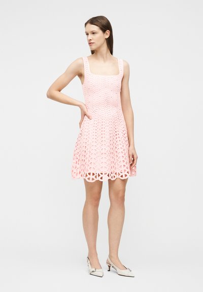 Γυναίκα στέκεται με το χέρι στη μέση, φορώντας ροζ δαντελένιο αμάνικο φόρεμα και λευκά μυτερά παπούτσια με χαμηλό τακούνι, πάνω σε απλό φόντο.